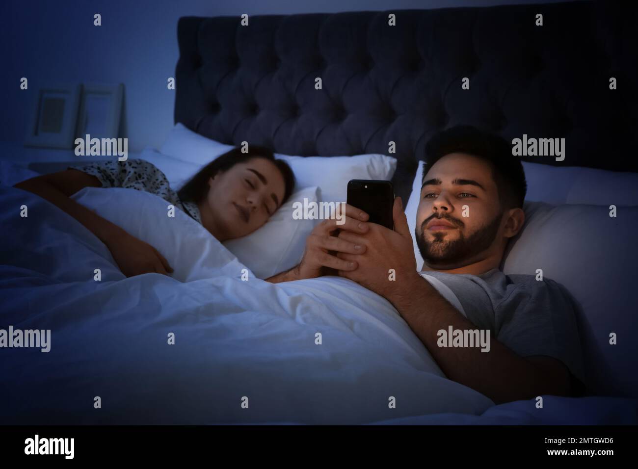 Young man using smartphone while his girlfriend sleeping in bed at
