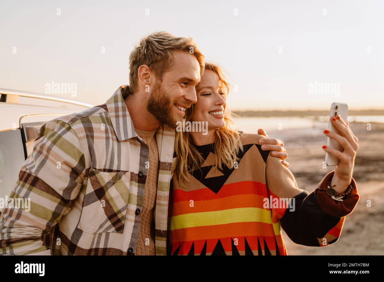 Happy young white couple taking selfie photo by car while walking at seashore on sunny day Stock Photo