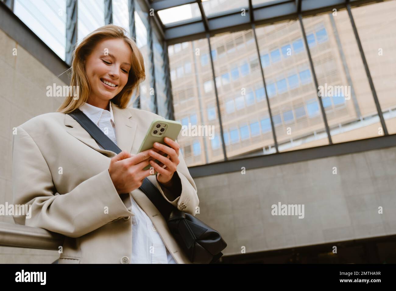 Young beautiful smiling happy woman holding and using her phone, while standing indoors leaning on the railing Stock Photo
