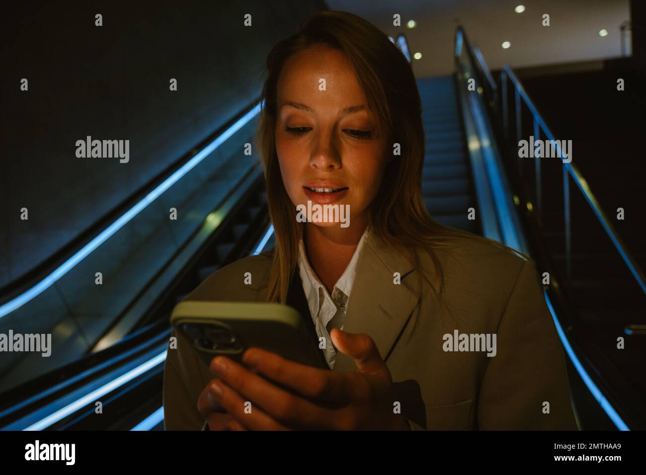 Portrait of young focused woman holding and using her phone, while standing in darkness on the escalator Stock Photo