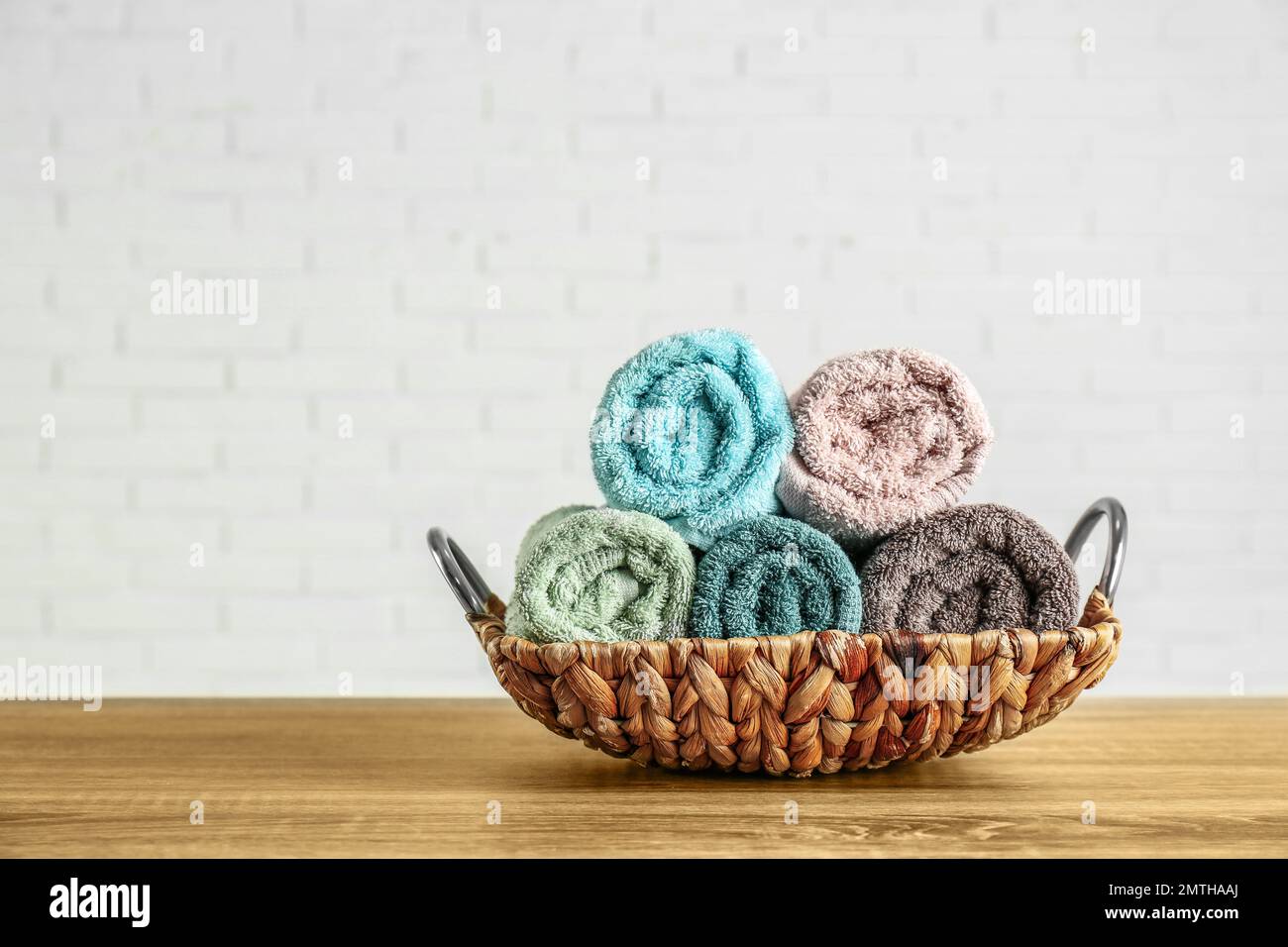 Wicker basket with rolled bath towels on wooden table near white brick
