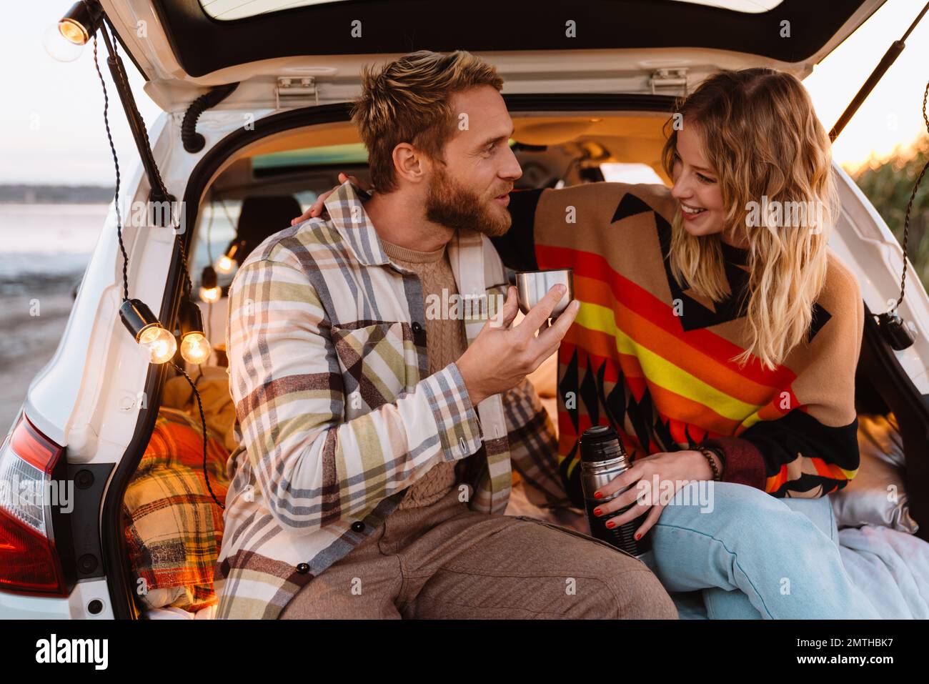 Happy young white couple smiling and drinking tea while sitting in trunk by seaside on sunset Stock Photo
