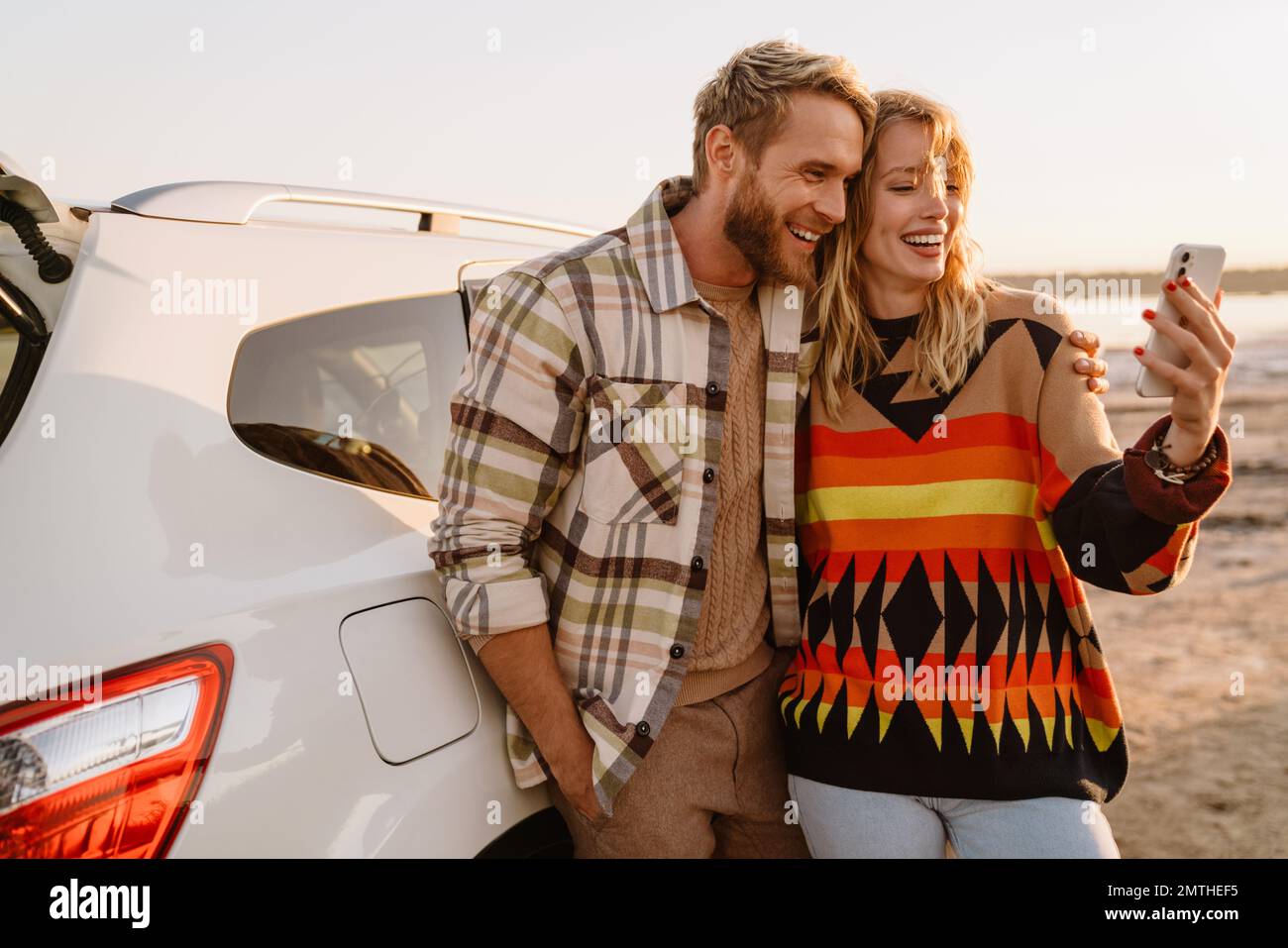 Happy young white couple taking selfie photo by car while walking at seashore on sunny day Stock Photo