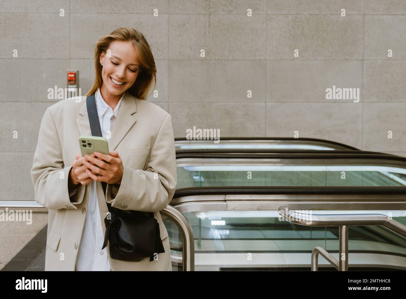 Young white woman smiling and using cellphone while standing by escalator outdoors Stock Photo