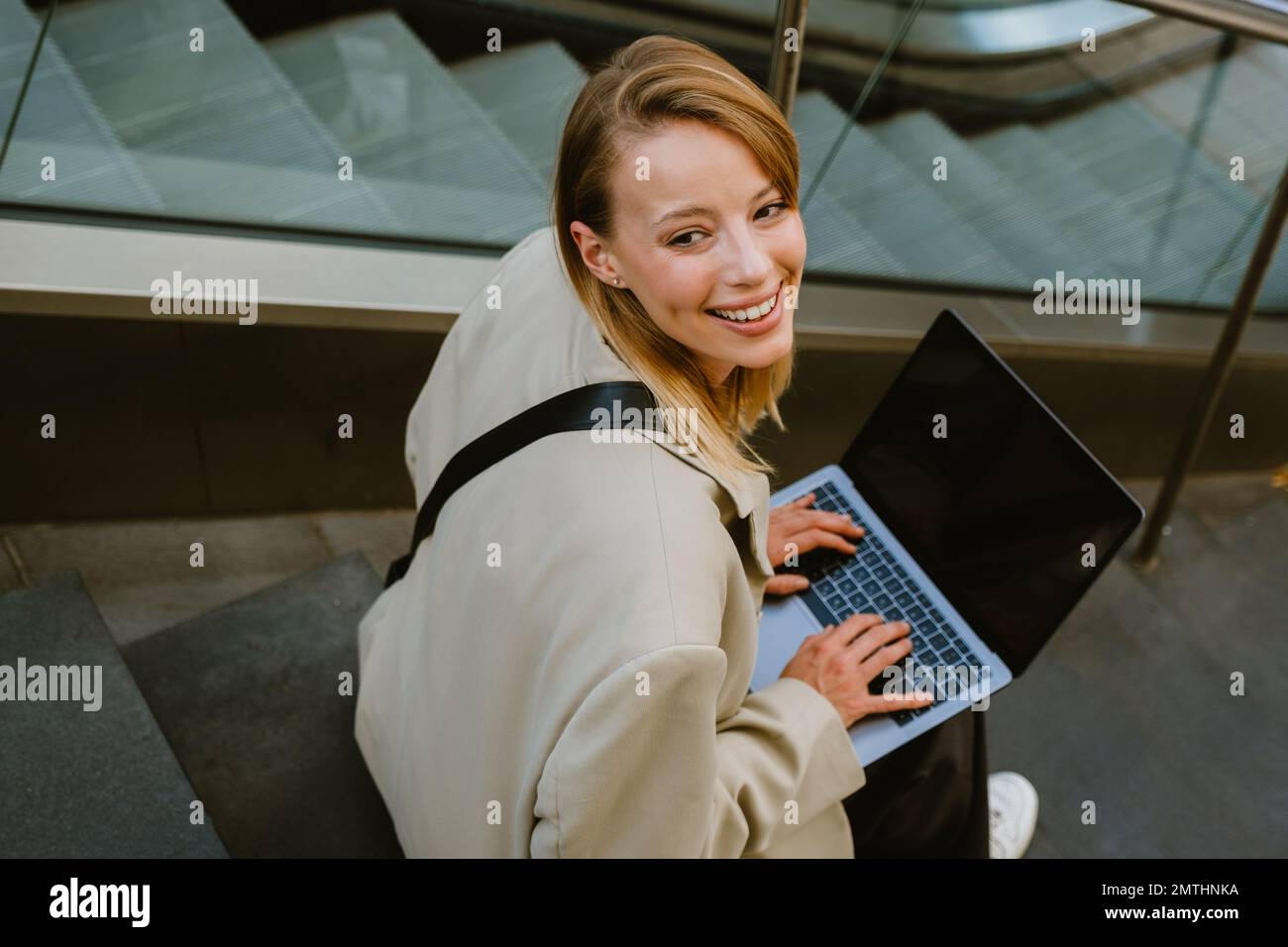 Young white woman smiling and using laptop while sitting on stairs outdoors Stock Photo