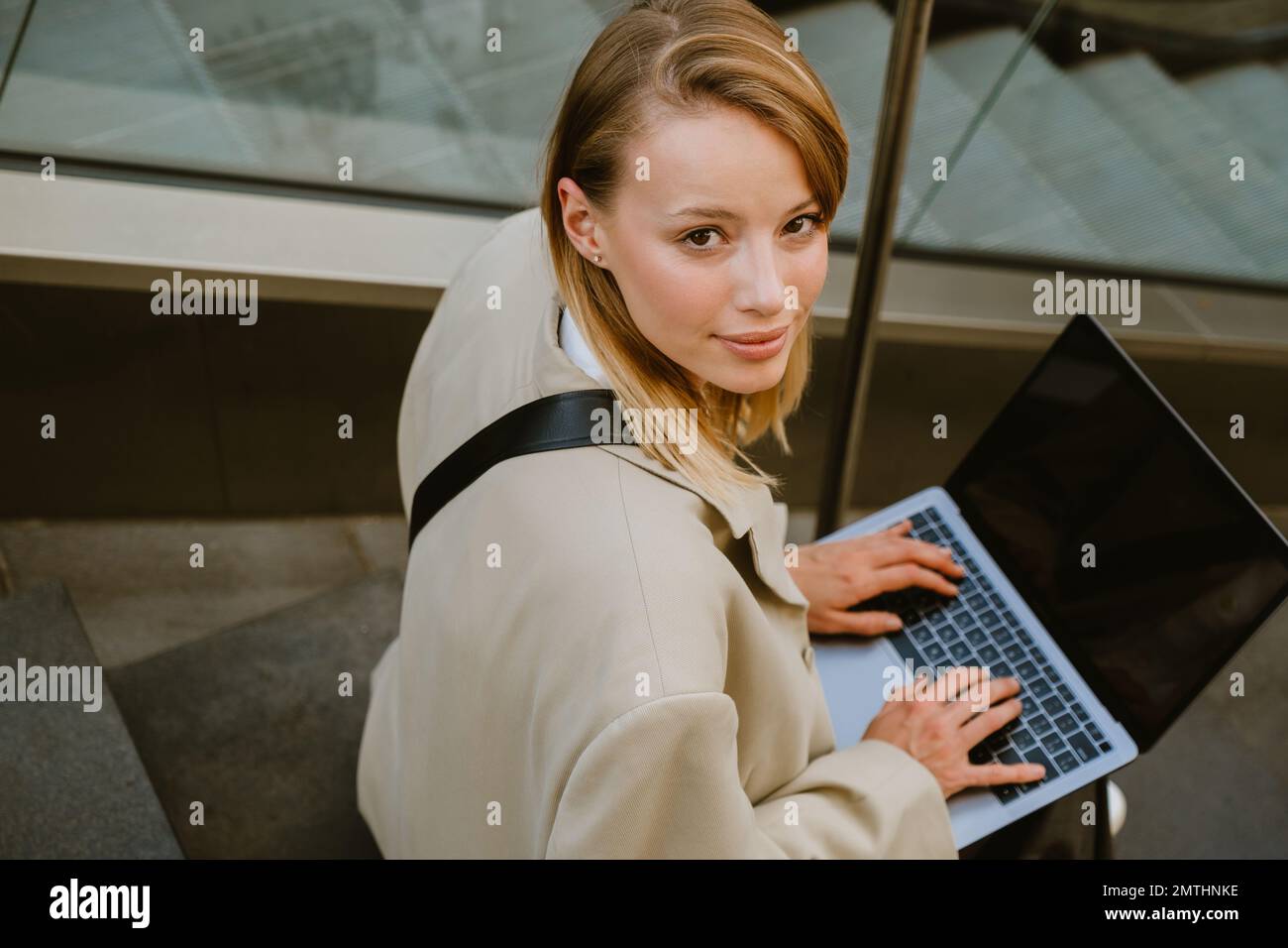 Young white woman smiling and using laptop while sitting on stairs outdoors Stock Photo