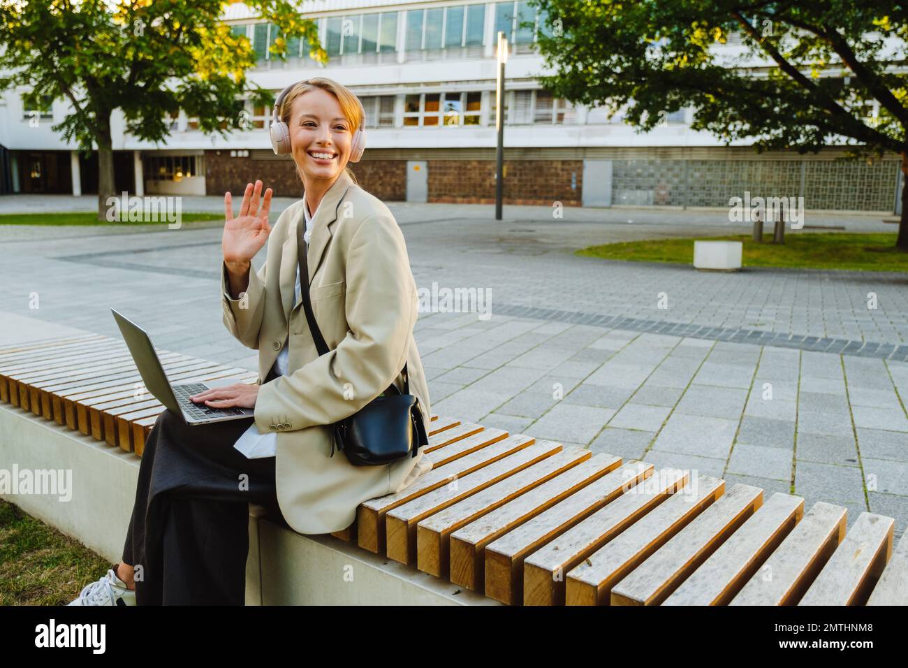 Young white woman using laptop and headphones while sitting on bench outdoors Stock Photo
