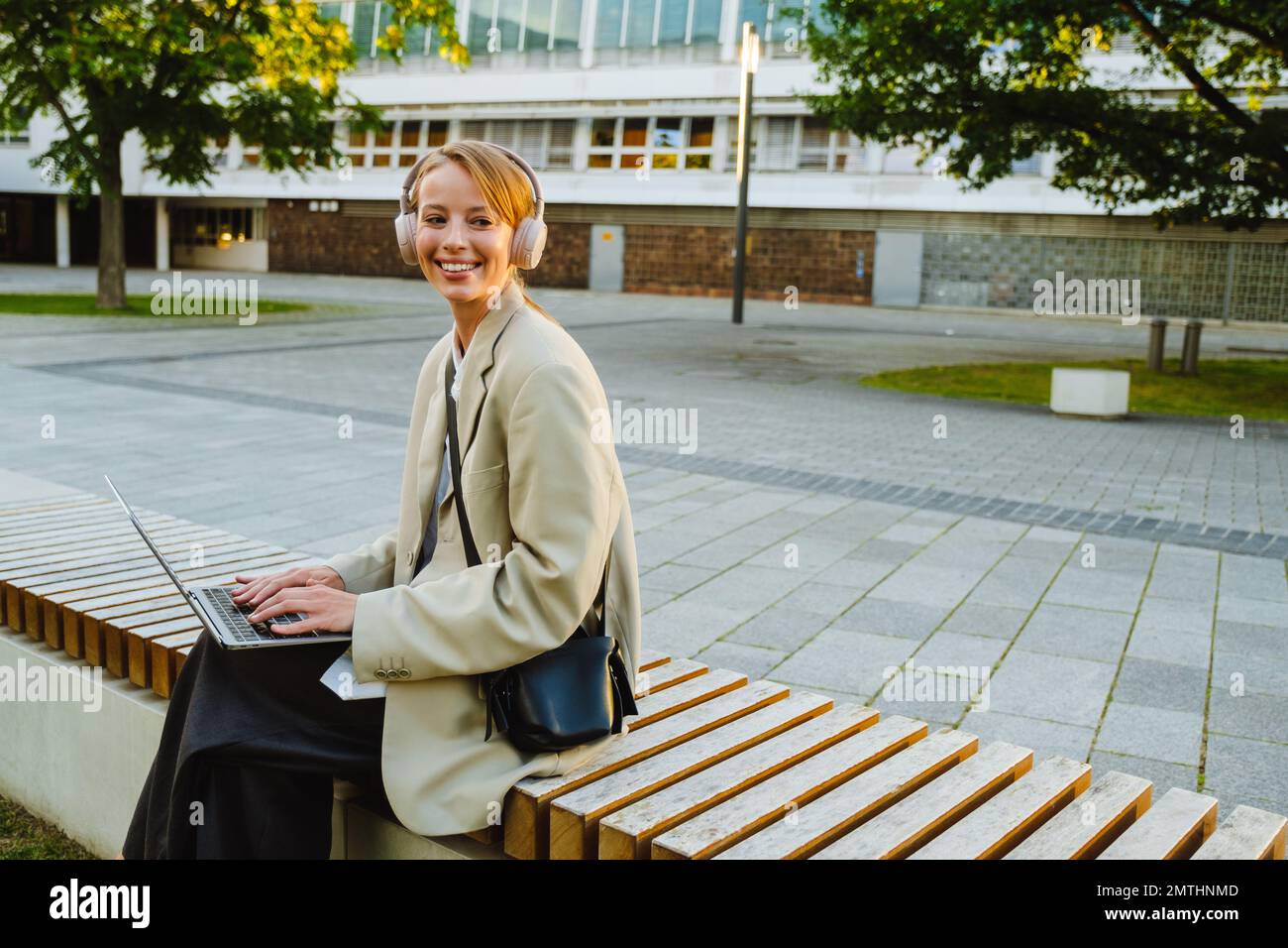 Young white woman using laptop and headphones while sitting on bench outdoors Stock Photo