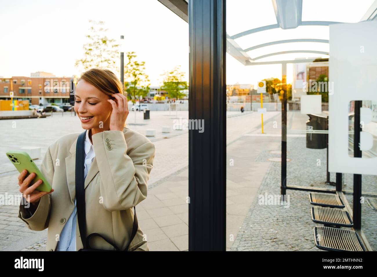Young white woman smiling and using cellphone while standing at bus station Stock Photo