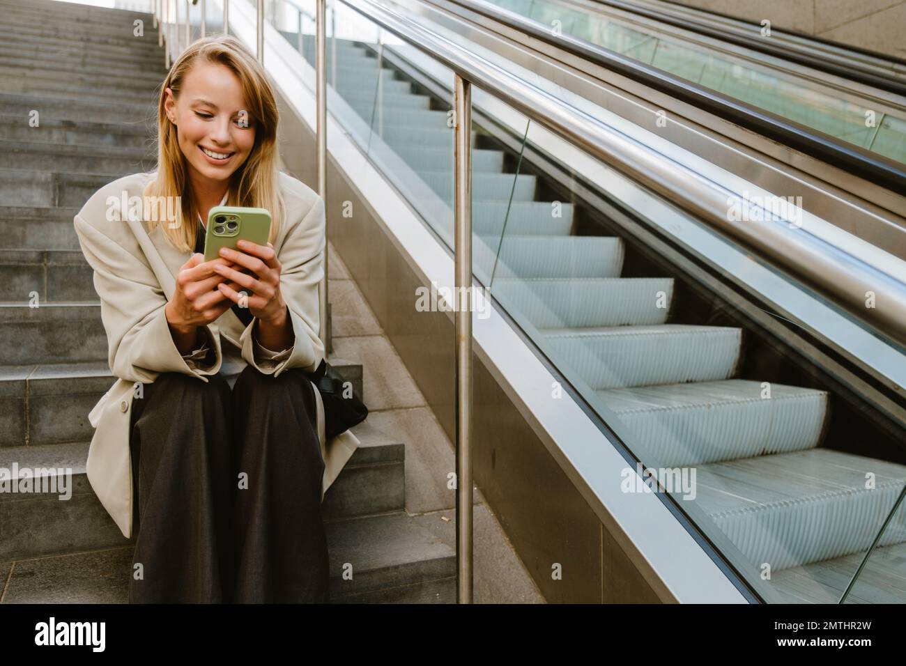 Young white woman smiling and using cellphone while sitting on stairs outdoors Stock Photo