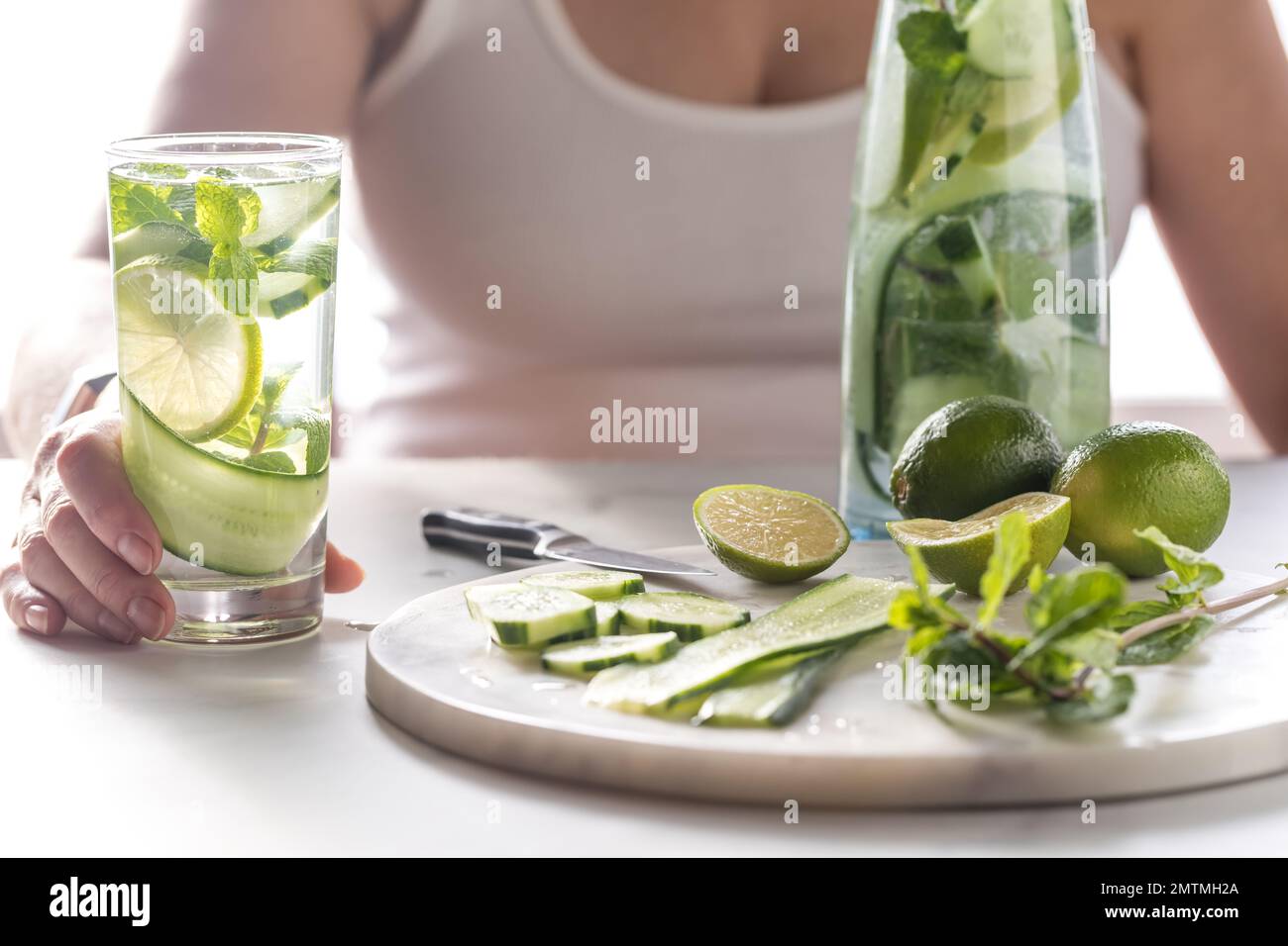Woman holding a glass of cucumber water with ingredients on a marble slab. Stock Photo
