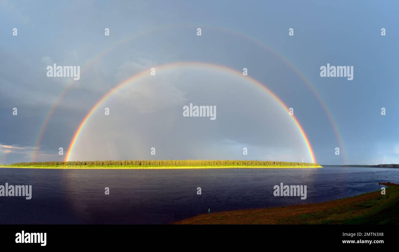 A fisher girl stands on the banks of the Vilyui River with a large double rainbow against the backdrop of a forest and a blue sky in Yakutia. Stock Photo