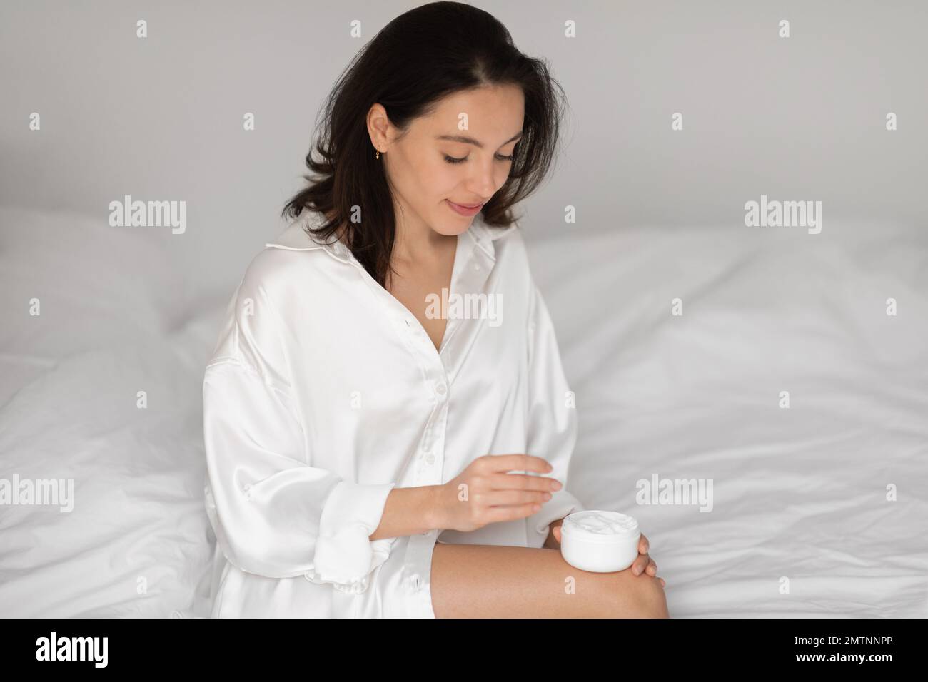 Smiling young european female in white silk robe sit on bed holding jar of cream, enjoy daily procedures Stock Photo