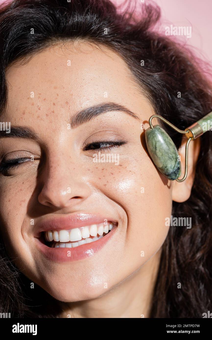 Close up view of smiling freckled woman using jade roller isolated on pink,stock image Stock Photo