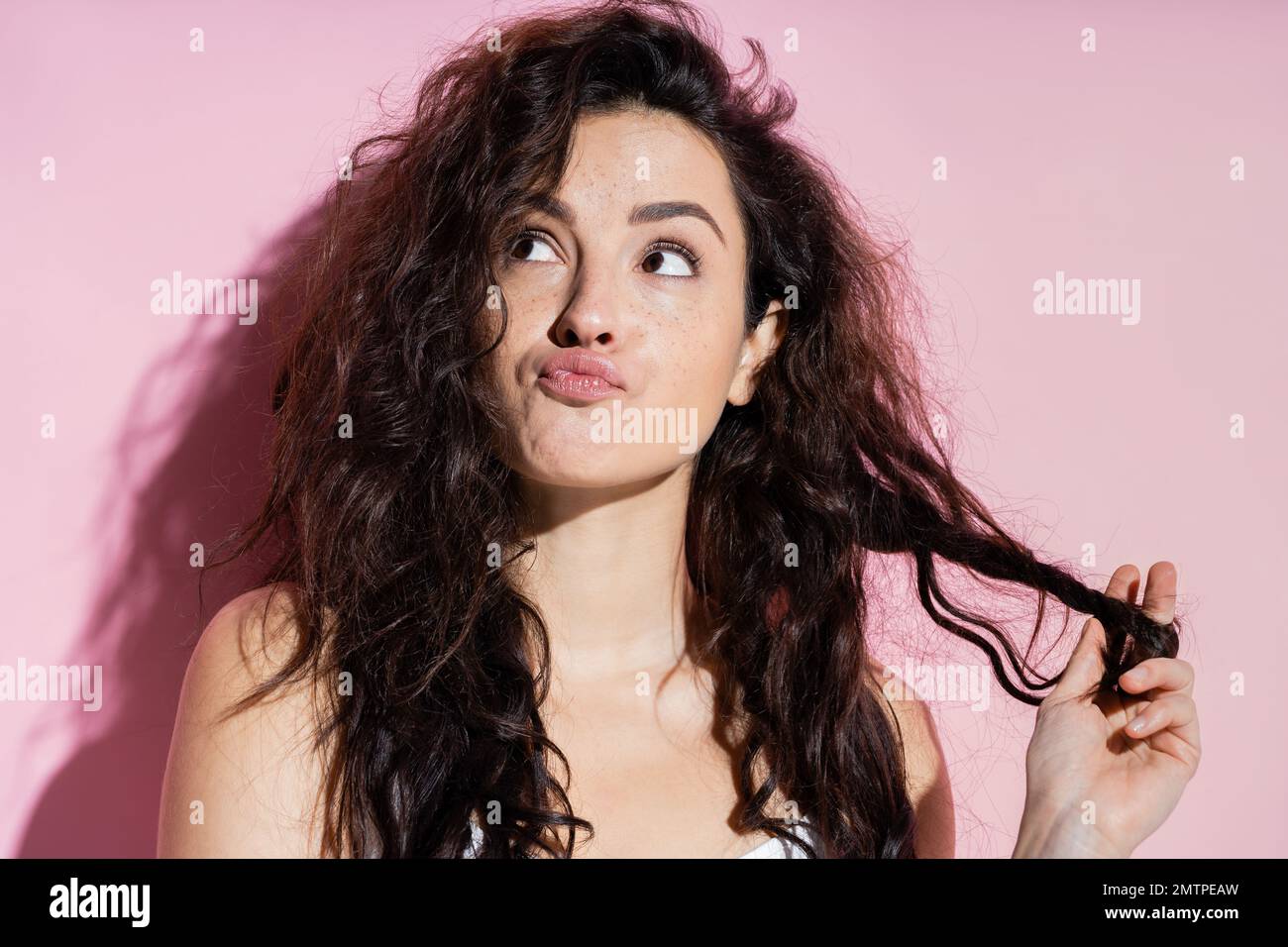 Pensive woman touching hair and pouting lips on pink background,stock image Stock Photo