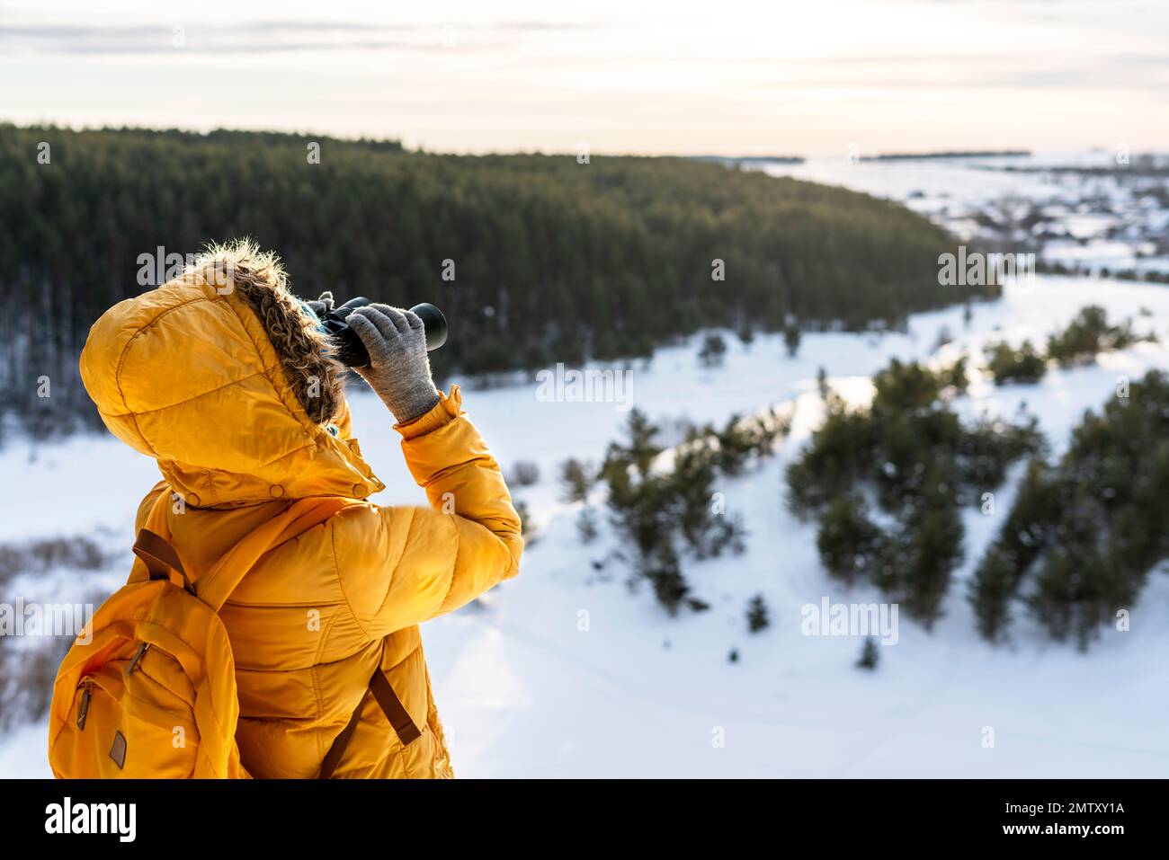 Young woman in yellow looking through binoculars at birds on snowy river against winter forest Birdwatching, zoology, ecology Research in nature, obse Stock Photo