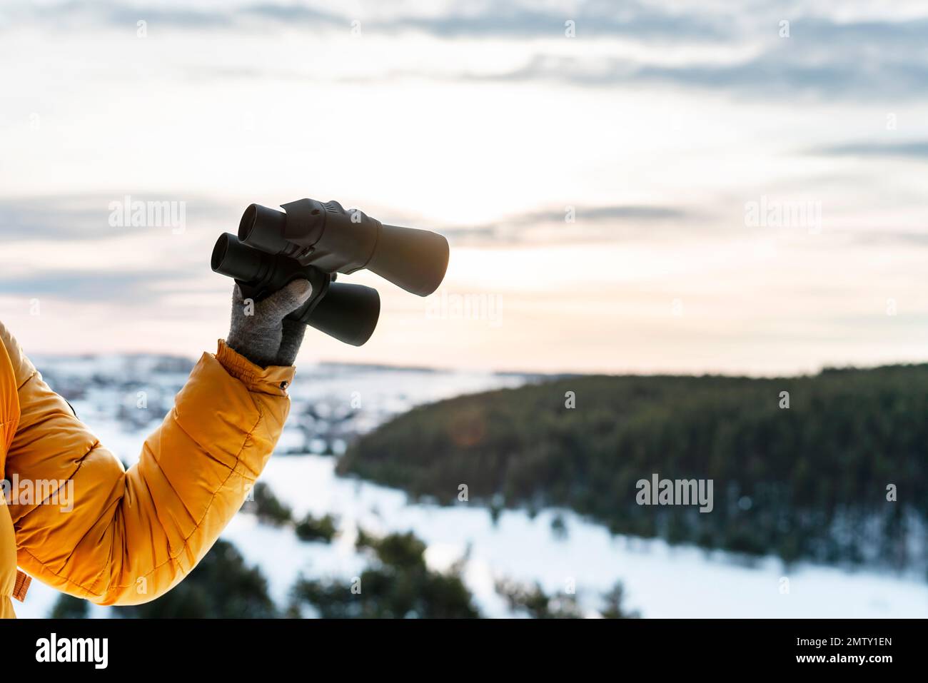 human hand in yellow jacket and warm glove holding binoculars againstwinter forest view of snowy river and sky Birdwatching ecology Research in nature Stock Photo