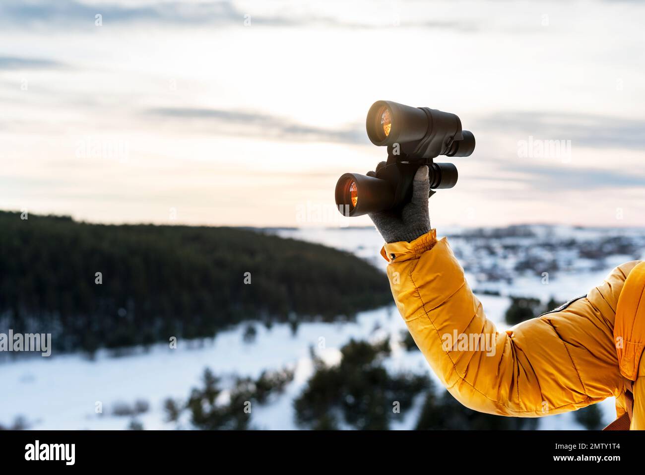 human hand in yellow jacket and warm glove holding binoculars againstwinter forest view of snowy river and sky Birdwatching ecology Research in nature Stock Photo