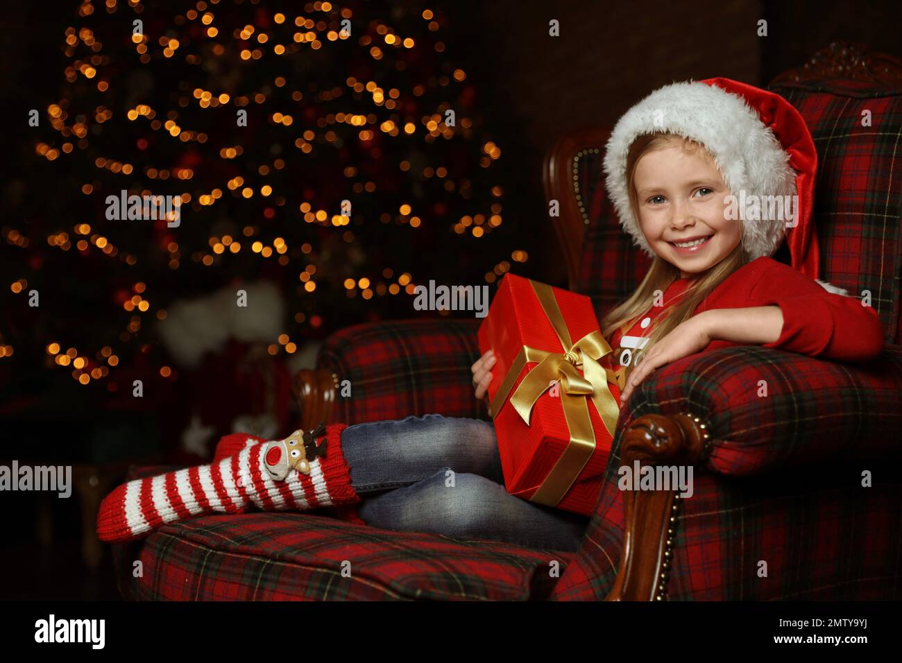 Cute little child with Christmas gift sitting in armchair at home Stock Photo