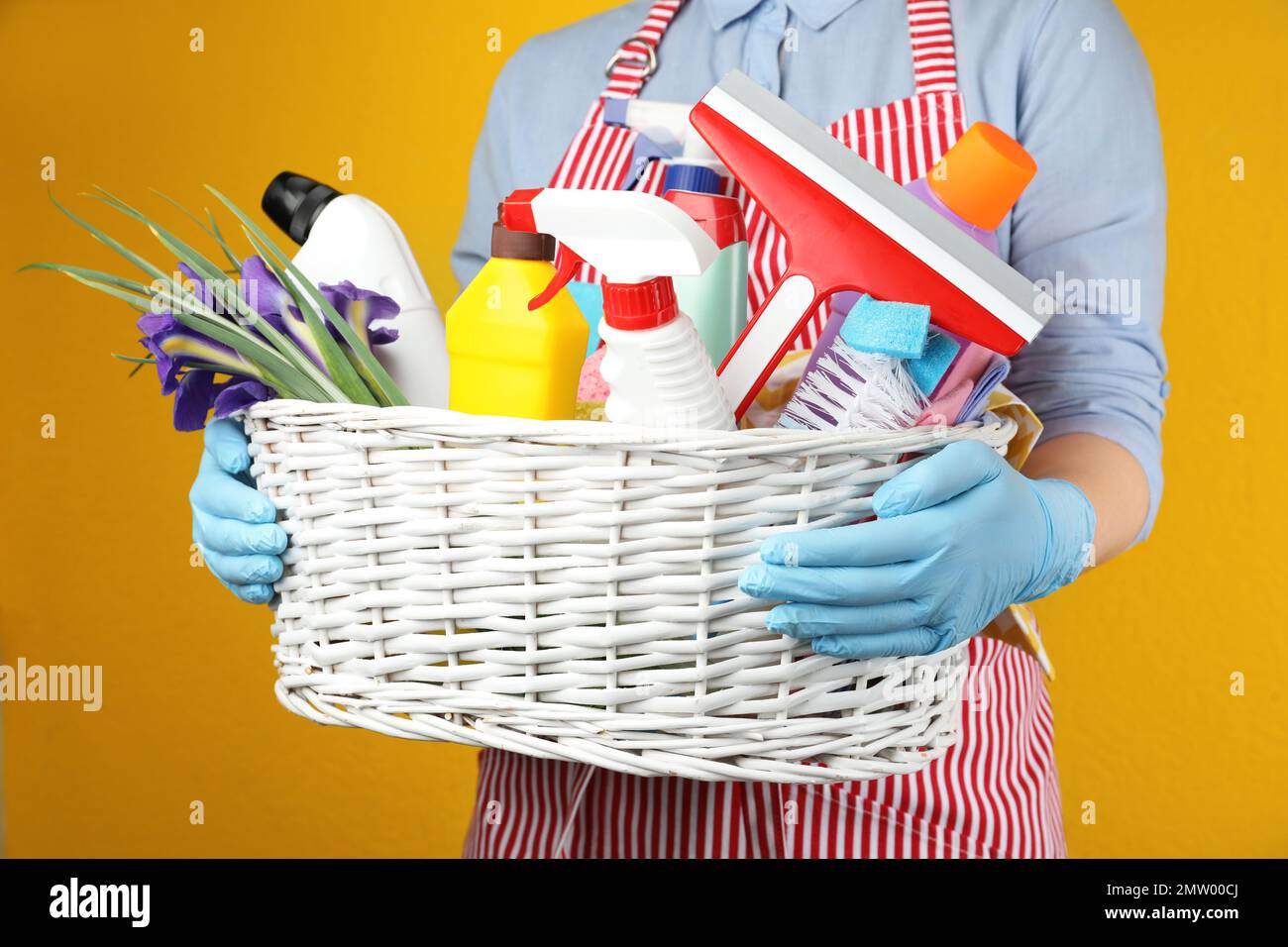 Woman holding basket with spring flowers and cleaning supplies on yellow background, closeup Stock Photo