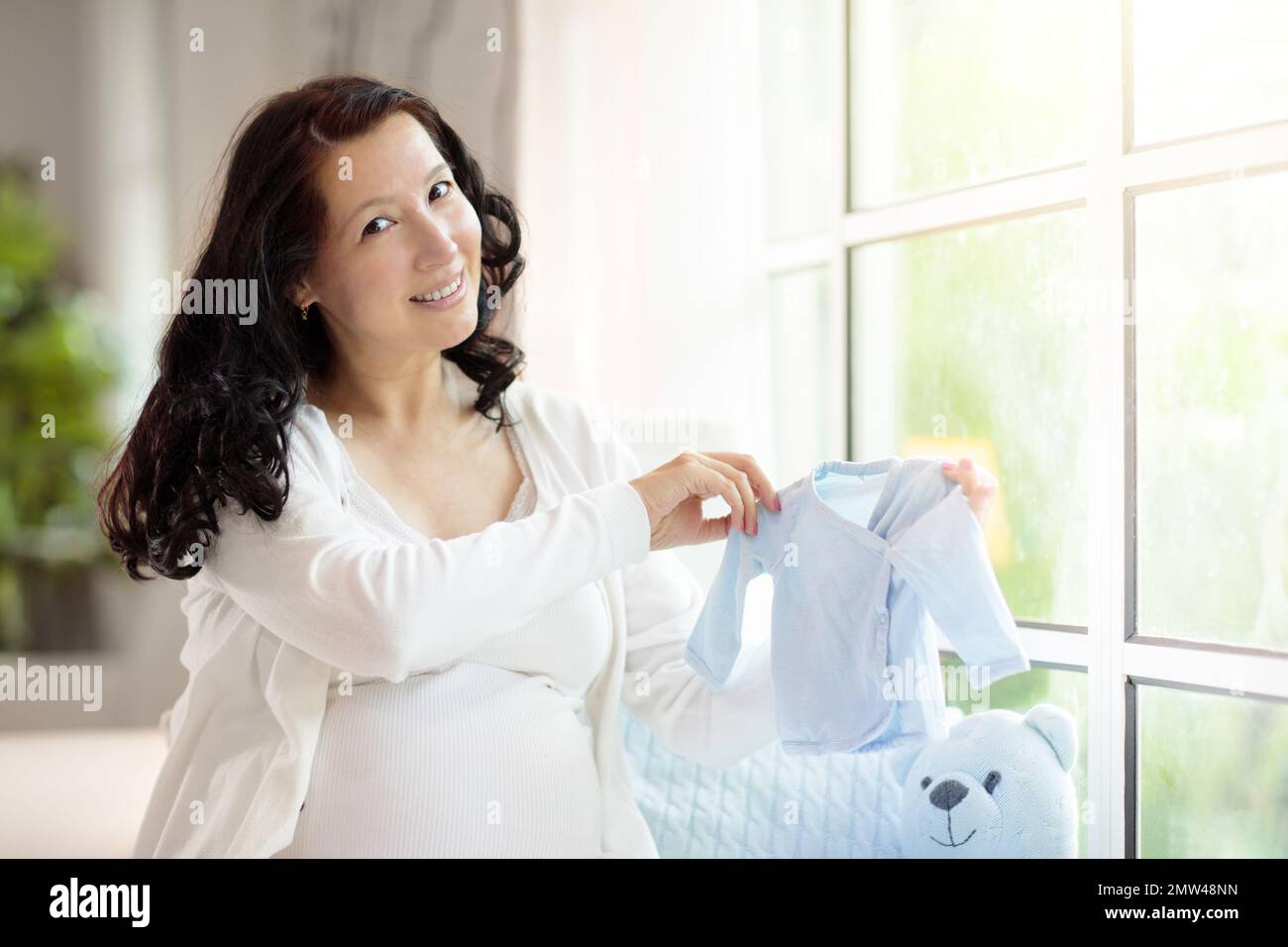 Pregnant woman at home. Asian expecting mother in white bedroom. Healthy pregnancy. Young female looking at baby sonogram preparing for birth. Materni Stock Photo