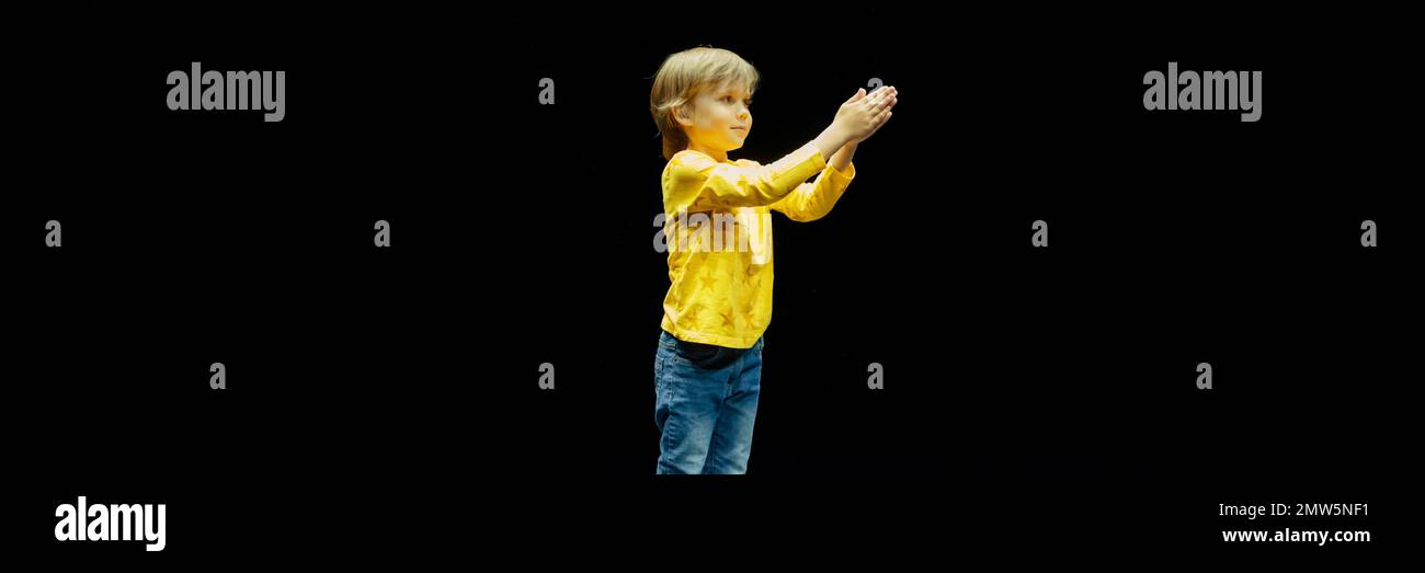 cute happy candid caucasian little six year old kid boy with long blonde hair stands tall full height and clap his hands in the studio illuminated by Stock Photo