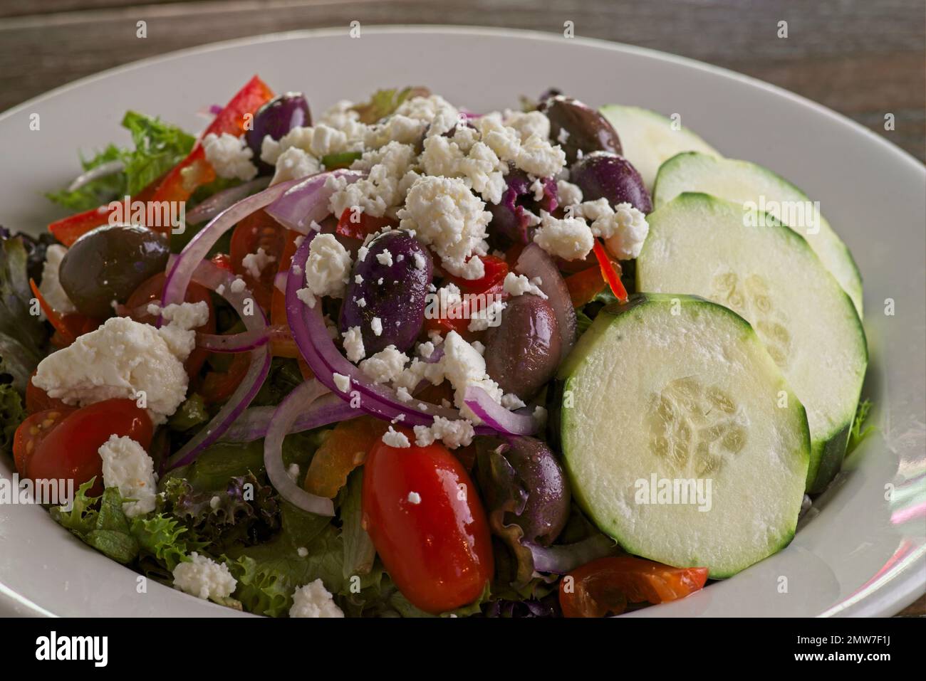 Greek salad with cucumber, cherry tomatoes, feta cheese, red onions, kalamata olives, and crisp lettuce, in a white bowl with dramatic side lighting Stock Photo