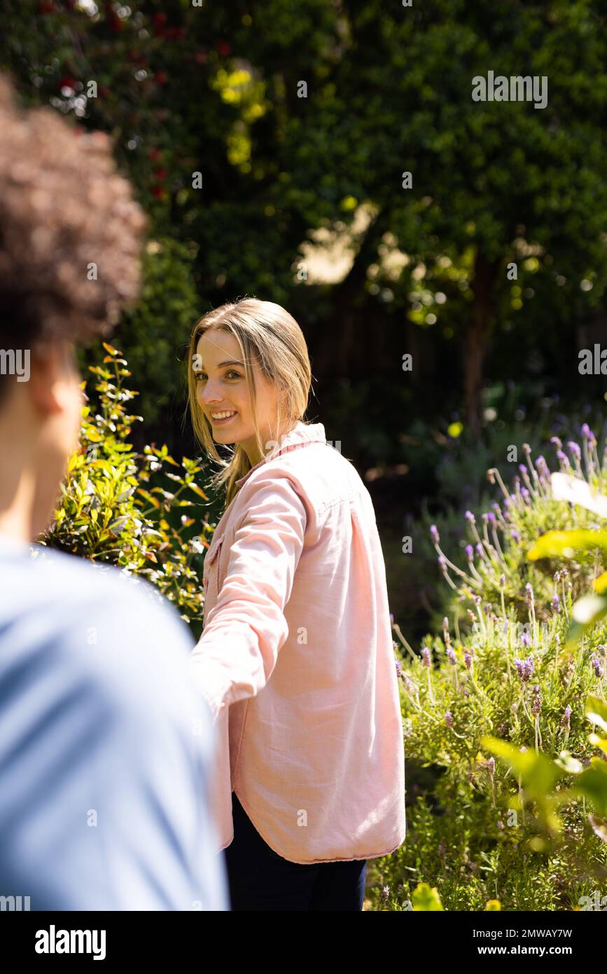Vertical of smiling caucasian woman leading diverse partner through sunny garden, with copy space Stock Photo