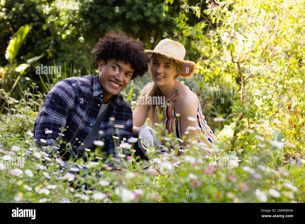 Portrait of happy diverse couple gardening, sitting amongst flowers in sunny garden Stock Photo