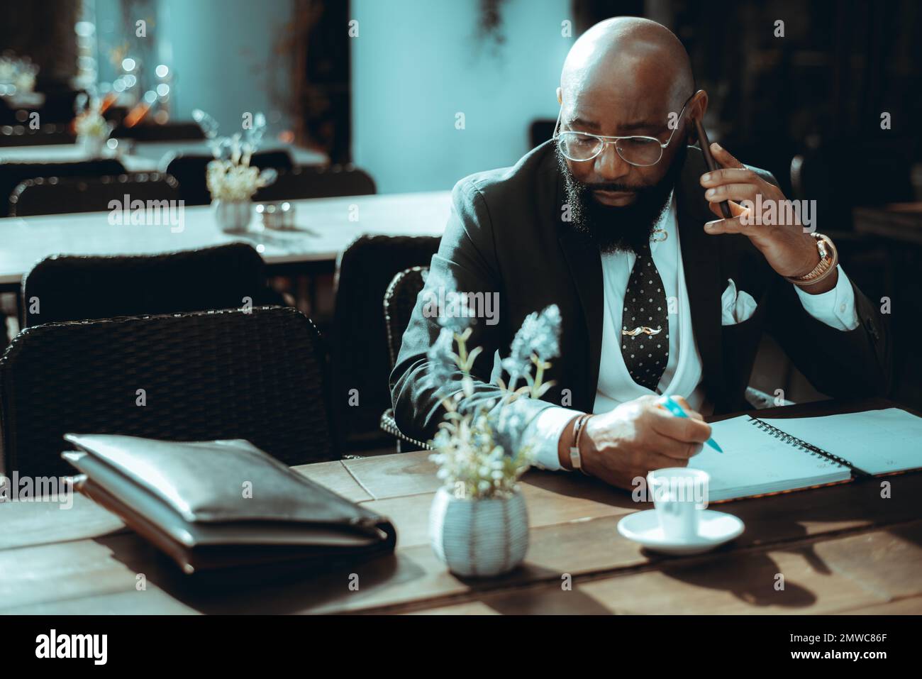A Black businessman with a beard sits at an outdoor cafe, dressed in a dark suit, a necktie, and a white vest writes notes in a notebook while talking Stock Photo