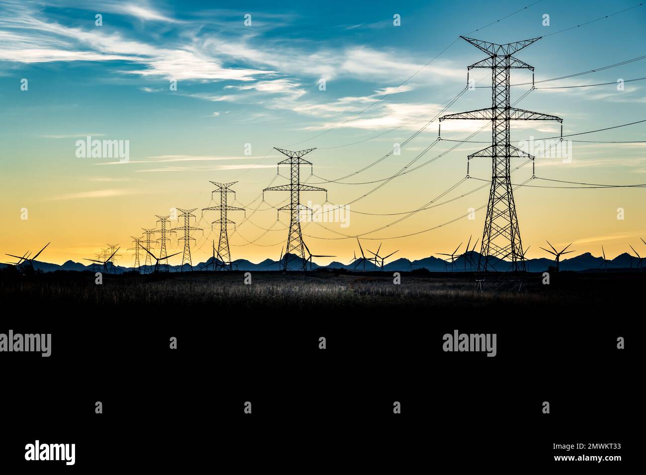 Sunset silhouette of steel lattice transmission towers and power lines overlooking distant mountains and windmills. Stock Photo