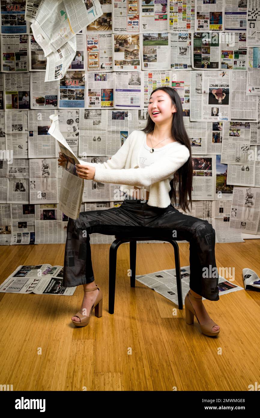 Full Body Tall Teenage Asian Girl Seated in Front of Backdrop of Newspapers Stock Photo