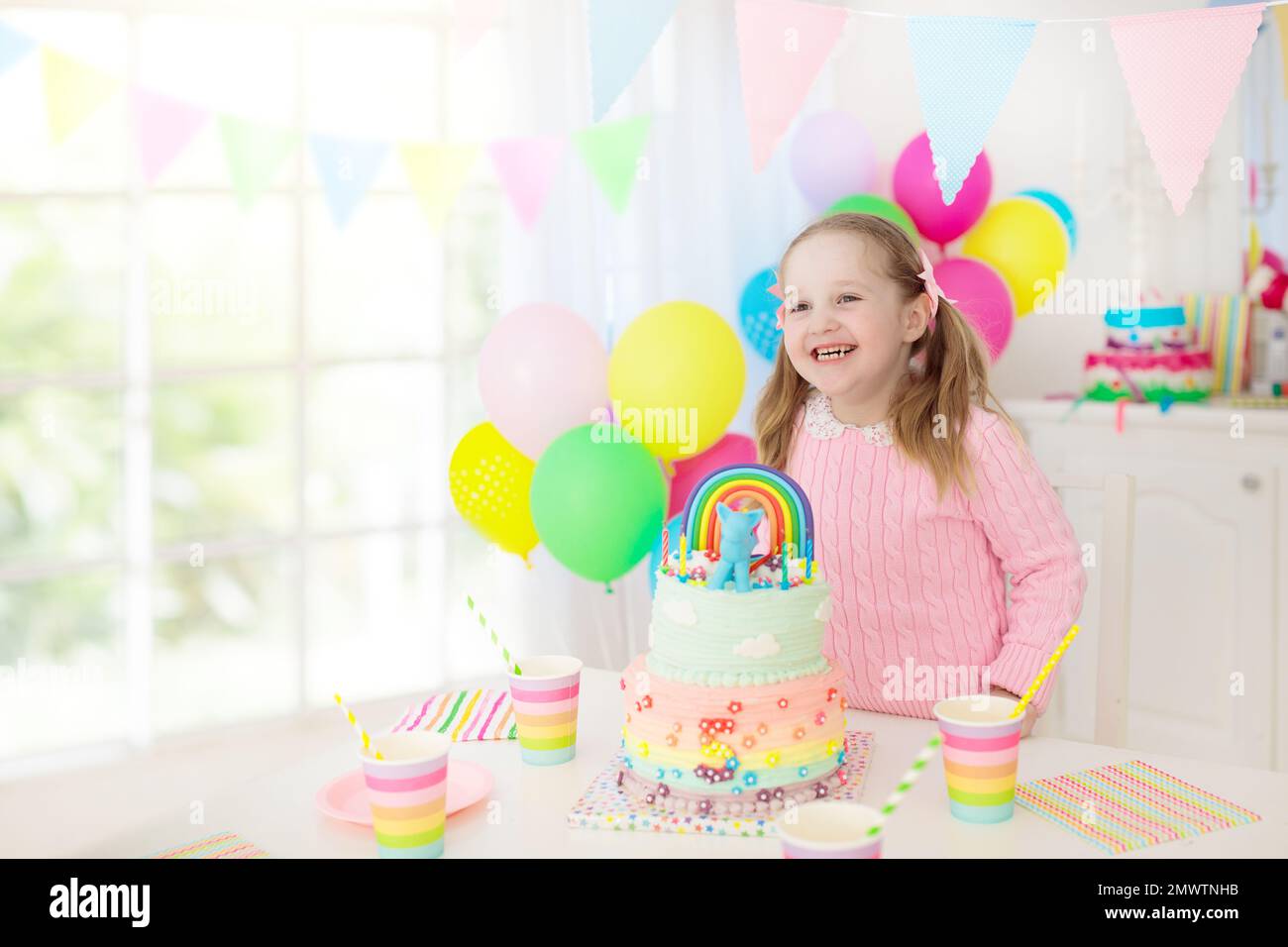 Kids birthday party with colorful pastel decoration and unicorn rainbow cake. Little girl with sweets, candy and fruit. Balloons and banner Stock Photo