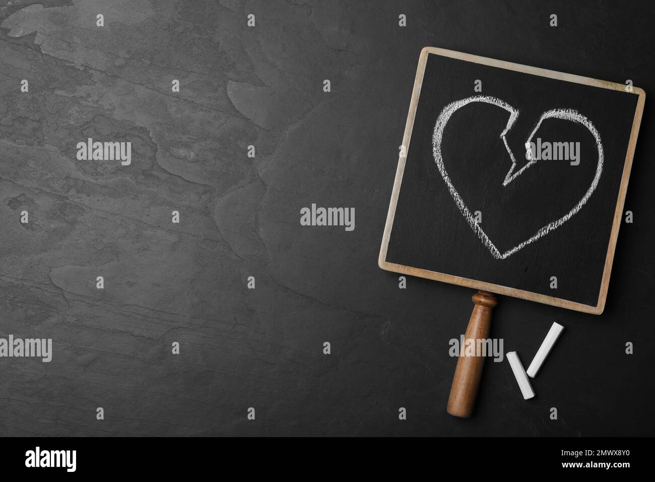 Blackboard with drawn broken heart and pieces of chalk on dark background, flat lay