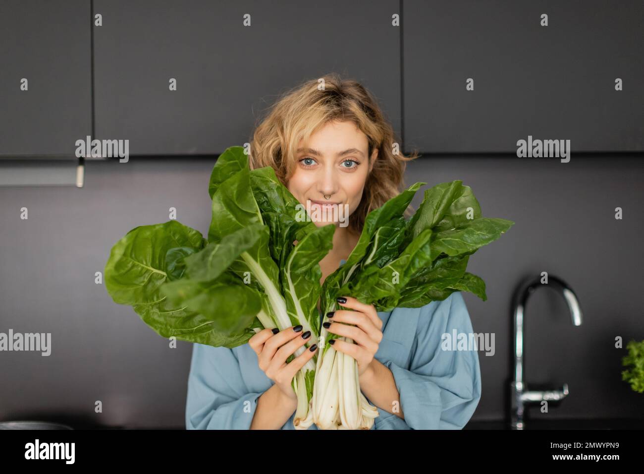pierced young woman smiling and holding green cabbage leaves in kitchen,stock image Stock Photo