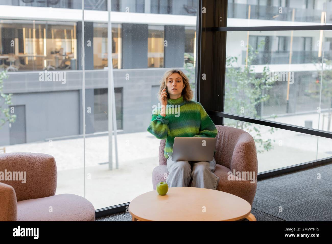 young woman in sweater talking on smartphone while sitting with laptop in lobby of hotel in Barcelona,stock image Stock Photo