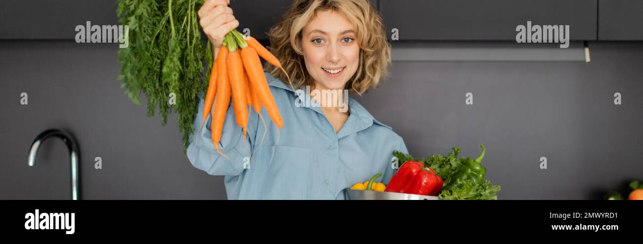 cheerful young woman holding bowl with vegetables and fresh carrots in kitchen, banner,stock image Stock Photo
