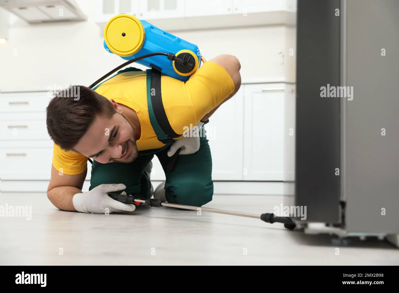 Pest control worker spraying insecticide under refrigerator in kitchen