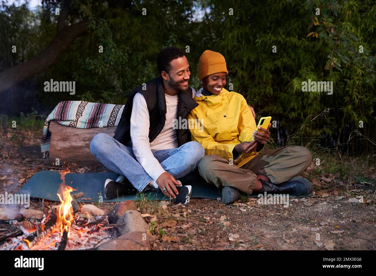 Cheerful couple sitting at camping in the mountain with campfire, using cellphone taking selfies. Stock Photo