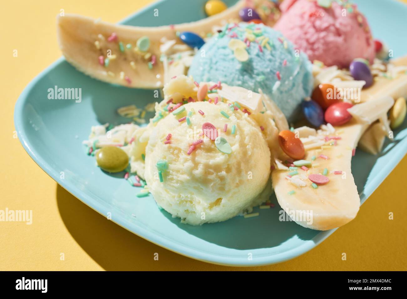 From above closeup of plate with banana split dessert with ice cream balls decorated with colorful dragee and sprinkles on yellow background Stock Photo