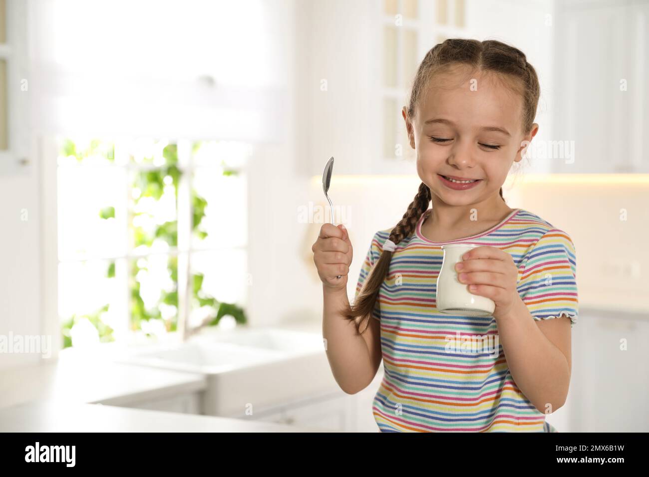 Cute little girl with tasty yogurt in kitchen. Space for text Stock