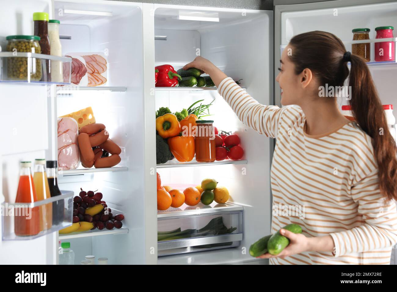 Young woman taking cucumber out of refrigerator indoors Stock Photo