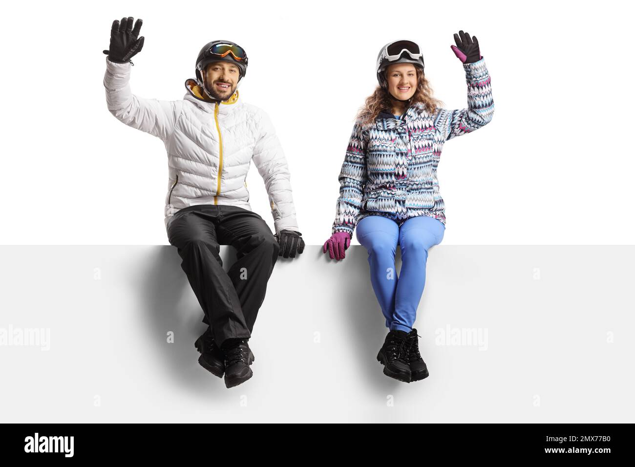 Young couple wearing winter skiing clothes sitting on a blank panel and waving isolated on white background Stock Photo