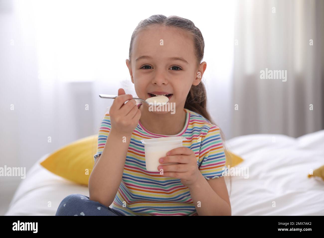 Cute little girl eating tasty yogurt on bed at home Stock Photo Alamy