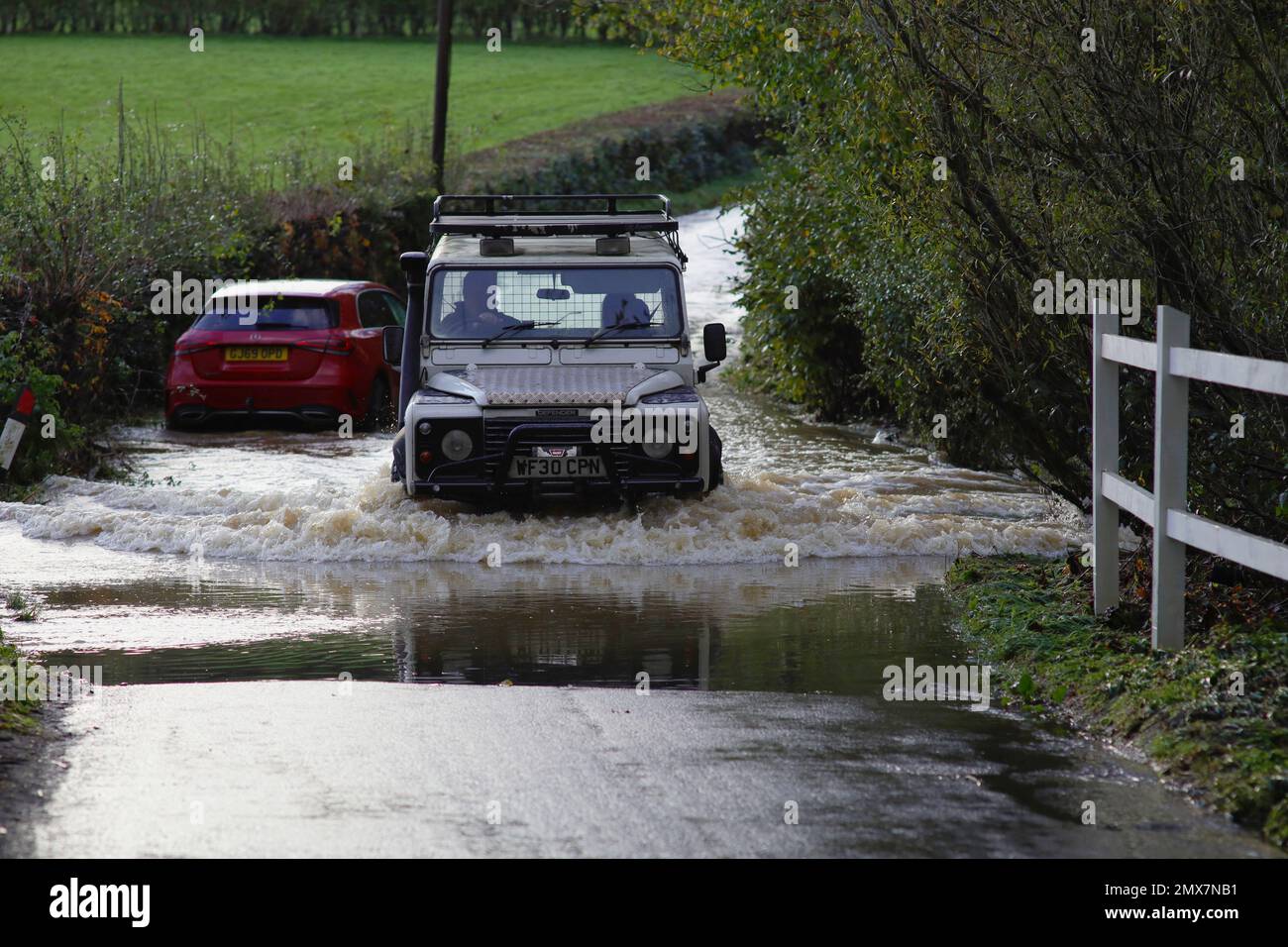 Land Rover Defender 110 passing through water from a swollen stream - tributary of the River Uck - which has flooded the England, East Sussex, lane between Buxted and Uckfield. Stranded Mercedes car in the background that had broken down in the flood water. Wealden Way crosses the road at this point. Stock Photo