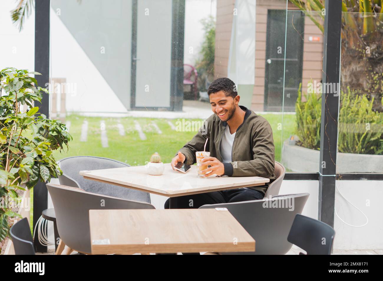 Optimistic ethnic male in casual outfit smiling and looking at screen while browsing smartphone and drinking beverage during break in earbuds in cafe Stock Photo