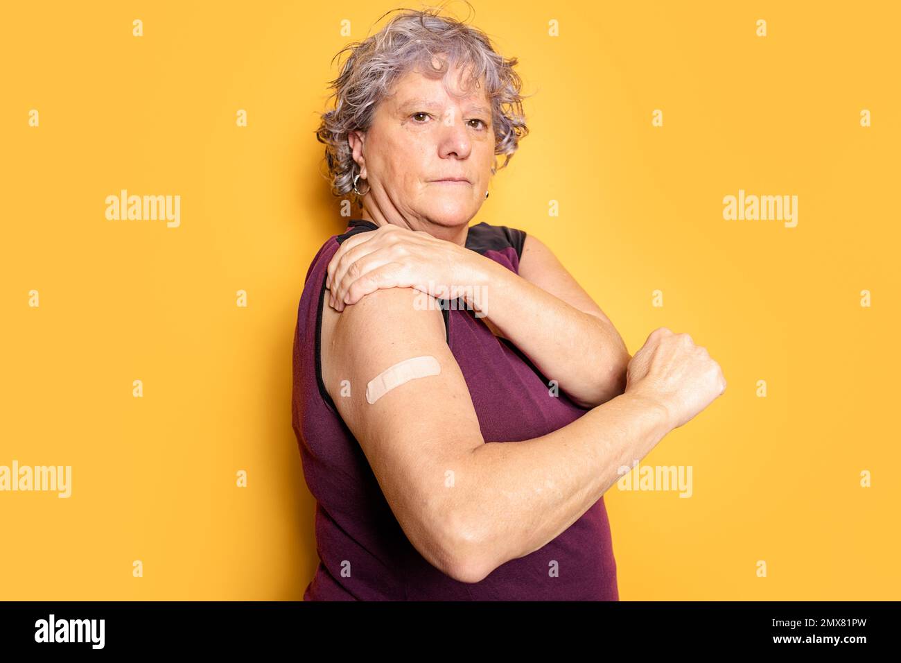 Serious senior female in t shirt looking at camera while demonstrating medical patch against yellow background Stock Photo