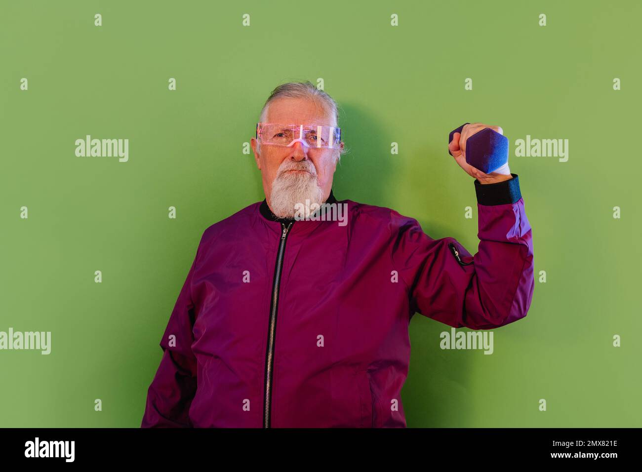 Serious bearded senior male hipster in casual clothes and glasses standing against green background while lifting dumbbell Stock Photo