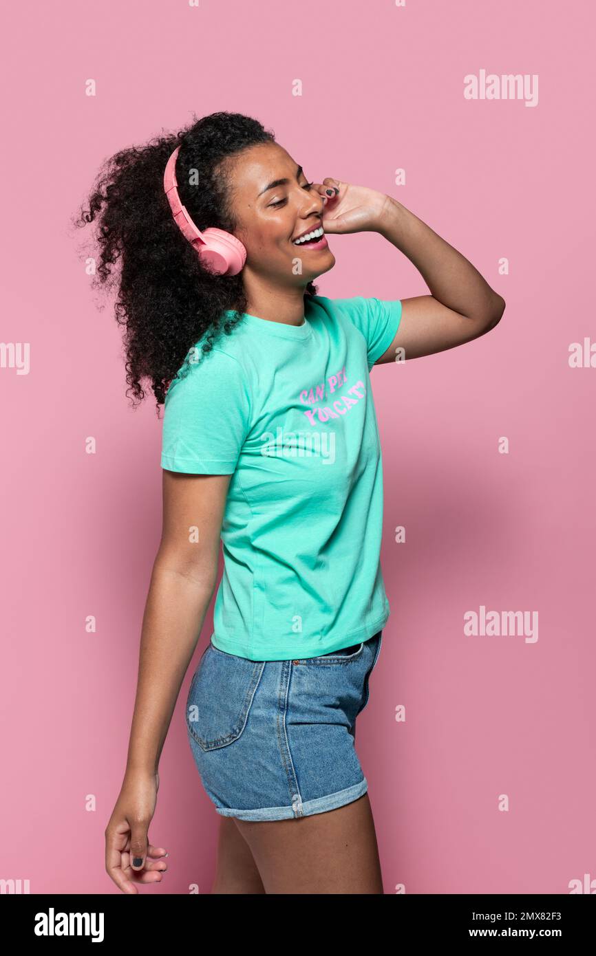 side view of happy young African American female in casual clothes with headphones while standing against pink background Stock Photo