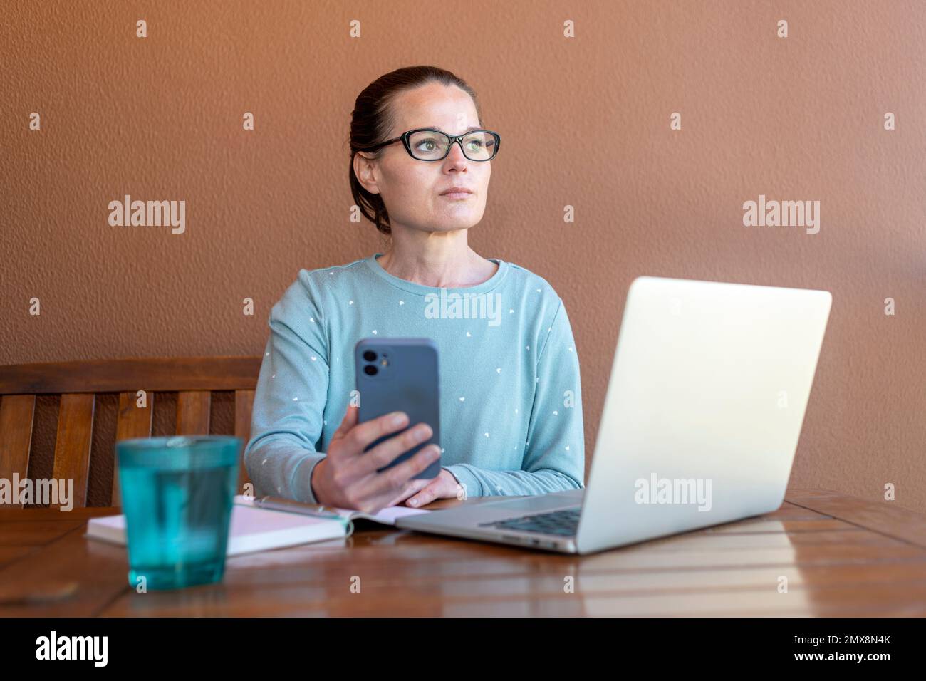 Woman sitting in her pyjamas working on her laptop and phone, looking away thinking. Stock Photo